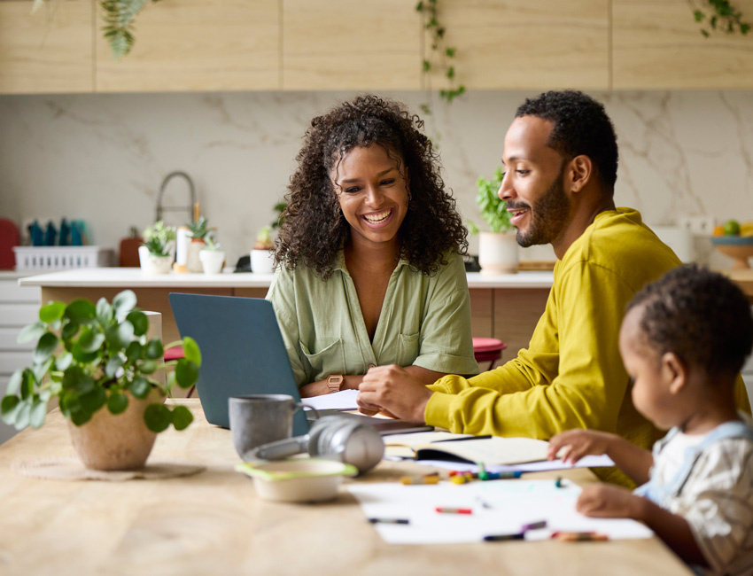 A smiling family sitting together at a kitchen table, with two adults looking at a laptop and a young child drawing nearby.