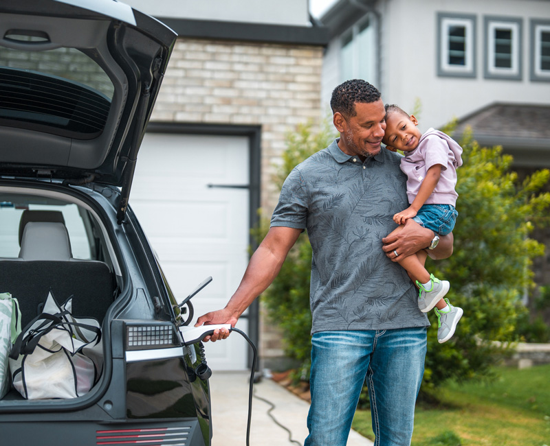Father holding a child while plugging in an electric vehicle at home in a residential driveway.