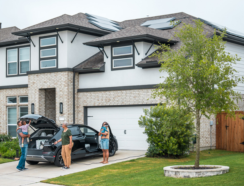 A family stands in the driveway of a modern two-story home with rooftop solar panels, next to a black electric vehicle being charged.